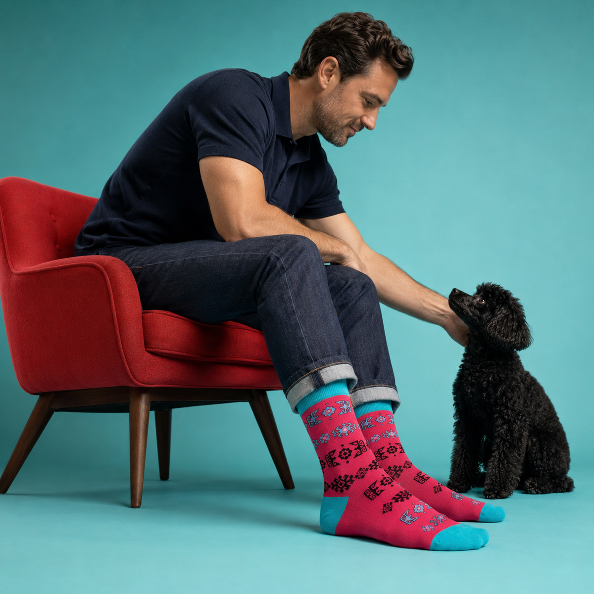 A man sitting in a red armchair next to a small black dog, wearing bright pink patterned crew socks with turquoise blue heels and toes, posed against a solid turquoise background.