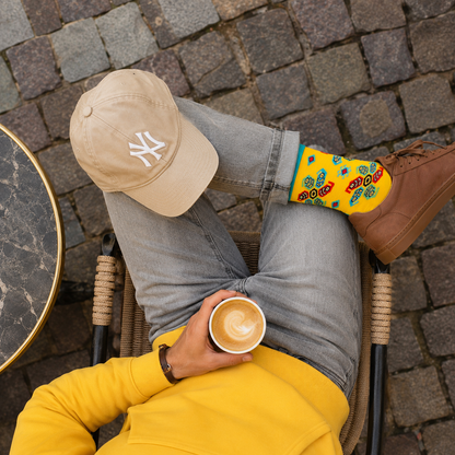 A person reclining outdoors on a woven chair over a cobblestone surface, wearing a beige New York Yankees cap, gray jeans, a bright yellow long-sleeve top, and yellow patterned designer cotton socks while holding a cup of coffee.