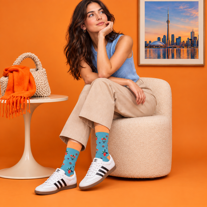 A woman sits on a beige upholstered chair in a bright orange room, wearing light blue ribbed tank top, beige cropped pants, white Adidas sneakers, and cotton crew socks with a red and yellow pattern. A white side table with a woven handbag and orange fringed throw sits to her left, and a framed print of Toronto hangs on the wall.