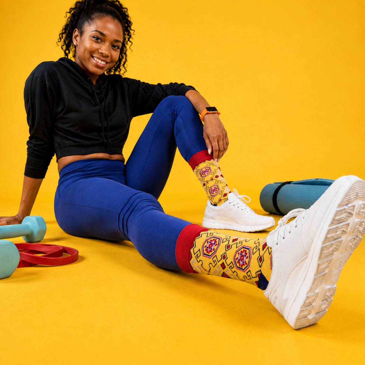 A woman sits on a bright yellow studio floor wearing a black cropped hoodie, blue leggings, white sneakers, and patterned crew socks. Yoga mats and dumbbells are visible around her.