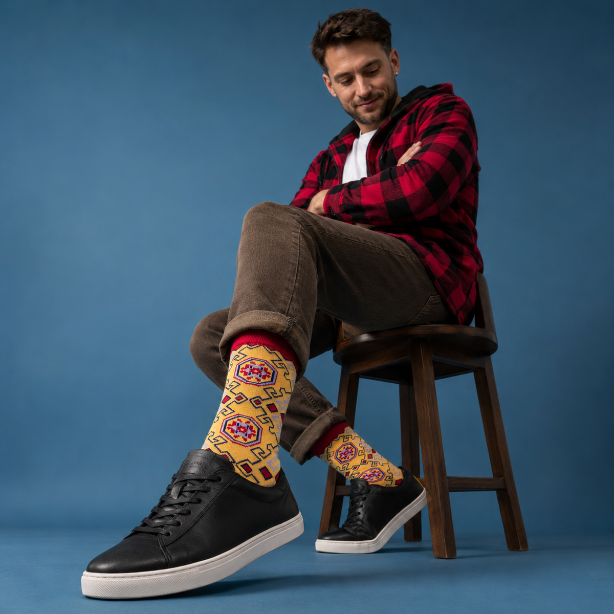A man sits on a wooden stool wearing a red and black plaid flannel shirt, brown pants, black sneakers, and distinctive yellow patterned crew socks against a solid blue studio background.