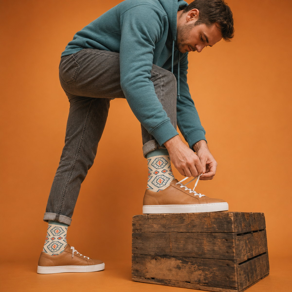 A man in a teal hoodie and gray jeans bends down to tie one of his tan low-top sneakers, while the other shoe is worn with a matching tan sneaker and a colorful geometric patterned sock. He is posed on a rustic wooden crate against a solid orange background.